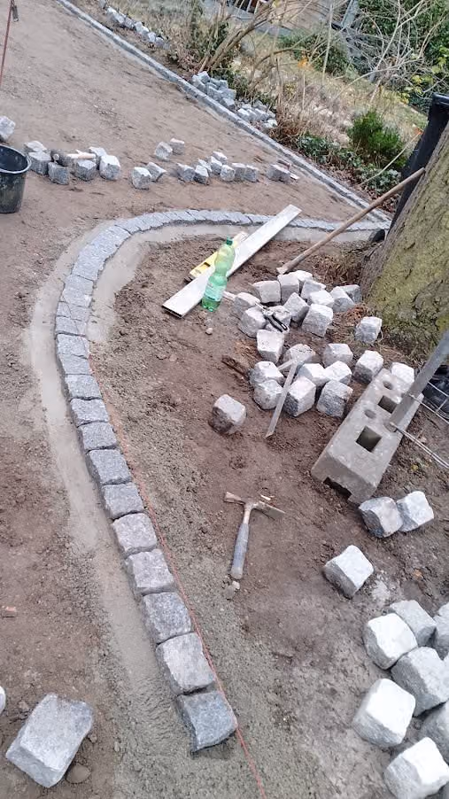 Geschwungene Rinne aus Granit-Kleinpflaster im Mörtelbett, Material daneben bereitgelegt
